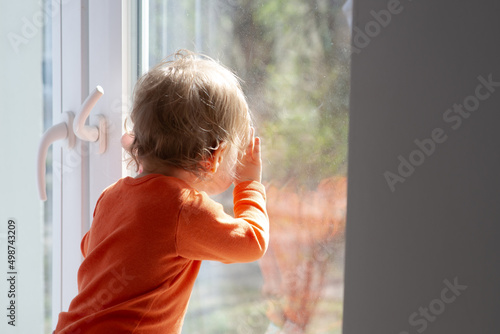 baby girl is looking through window glass at sunny day, touching the glass and having a fun with the sunbeams, natural dirty glass