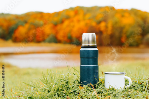 Camping thermos and enamel mug with hot drink and steam on shore near water against backdrop of colorful autumn forest in nature. Copy space, close-up