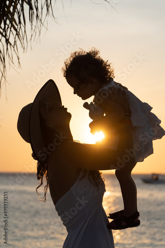 Young woman in white dress and sun hat holding in her arms her little baby daughter in ocean beach in Aruba island on sunset. High quality photo