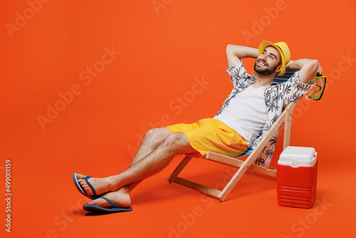 Young minded happy cool tourist man in beach shirt hat lie on deckchair near fridge hold hands behind neck isolated on plain orange background studio portrait Summer vacation sea rest sun tan concept