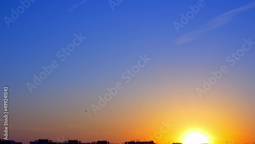 The setting sun over the roofs of city buildings in the distance is landing a passenger plane. Beautiful urban sunset. The blue sky is flooded with the yellow fire of the setting sun