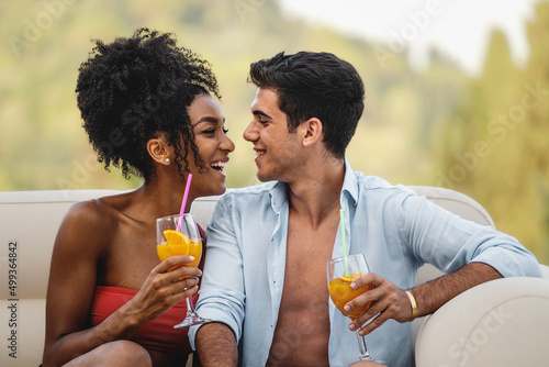 Happy biracial couple of young people sit outdoors in a sofa in the garden looking at each other while drinking healthy fruit cocktails - love and vacation lifestyle concept
