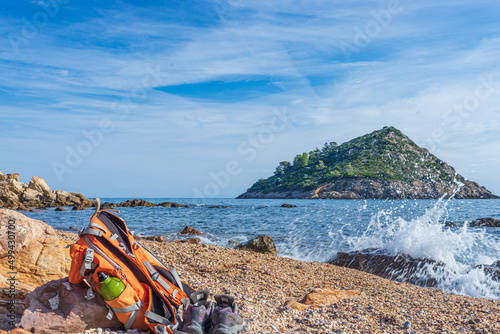 Backpack and hiking shoes on empty beach in Argentario region, Tuscany, Italy. Sand bay in natural park, island in mediterranean sea blue waving water