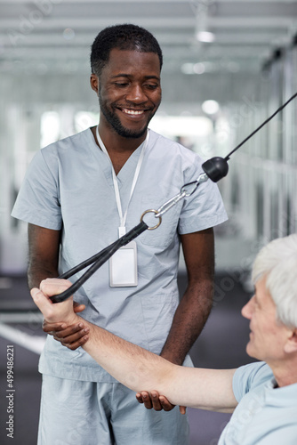 Vertical portrait of smiling rehabilitation therapist assisting senior man at gym in clinic