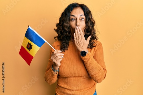 Middle age hispanic woman holding moldova flag covering mouth with hand, shocked and afraid for mistake. surprised expression