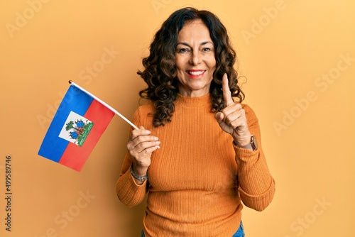 Middle age hispanic woman holding haiti flag smiling with an idea or question pointing finger with happy face, number one