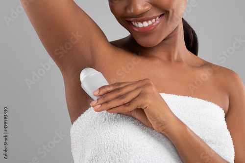 Satisfied young black female wrapped in towel applying deodorant to her armpit, isolated on gray background