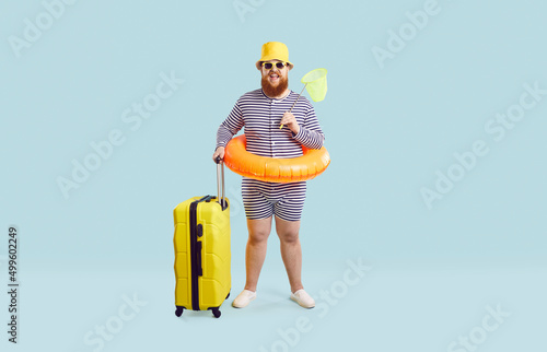 Full body length of happy funny fat man in striped swim suit, hat, glasses and floaty standing in studio and holding yellow suitcase and butterfly scoop net. Traveling vacation, summer holiday concept