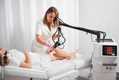 Dermatologist working with modern laser device for polishing and repairing skin defects. Beautician demonstrating work of CO2 laser, warming skin of clients thigh, stimulating local rejuvenation.