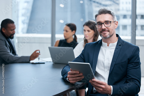 Were always busy making big things happen here. Portrait of a mature businessman using a digital tablet in an office with his colleagues in the background.