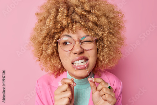 Dental health care concept. Beautiful young woman with curly bushy hair cleans teeth with dental floss wears transparent eyesglasses isolated over pink background shows how to floss correctly