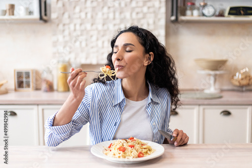 Young latin woman eating delicious pasta, enjoying tasty homemade lunch with closed eyes, sitting in kitchen
