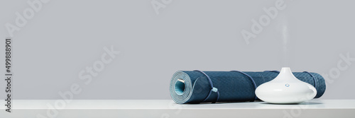 Rolled blue yoga mat and essential oil diffusor for enhanced practice. Ultrasonic and humidifying aromatherapy. Meditation fitness and exercise banner copy space. Active lifestyle. home or gym club