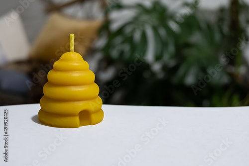 A beehive votive shaped beeswax candle is sitting on white paper with plants in the background.