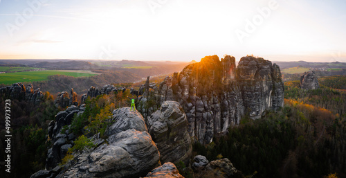 Male silhouette with raised arms on sharp mountain top. Sport life concept and outdoor activities. Illuminated rocks and forest by sun in the Saxon Switzerland, Germany