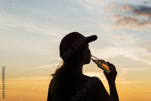 Silhouette of girl in baseball cap drinking soda water from glass bottle and looking at sunset sky. Rear view of female