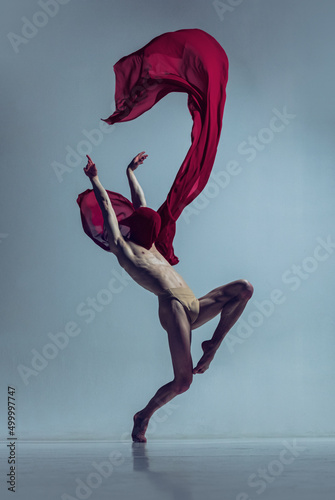 Portrait of young man, flexible ballet dancer in action with red fabric, cloth isolated on navy studio background. Grace, art, beauty concept.