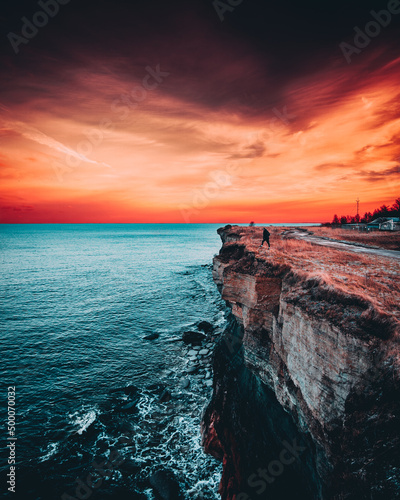 Vertical shot of a man on the edge of a cliff near the sea with turqoise blue water and red sunset