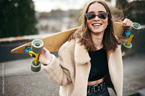 Stylish young woman holding skateboard on shoulders on roof