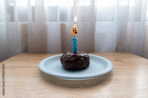 pequeño pastel muffin de chocolate en plato azul con vela azul encendida y flama de emoji sobre mesa de madera y cortinas blancas al fondo