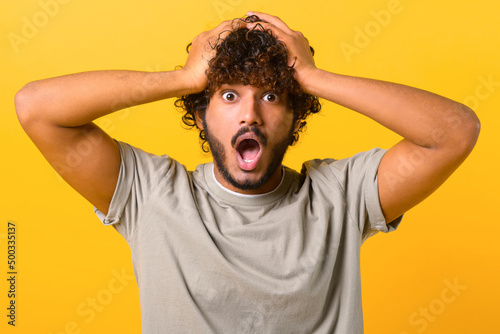 Excited shocked Indian man holding hands on head and screaming looking at camera with big eyes and open mouth, shocked amused with news, win at lottery. Indoor studio shot isolated on yellow
