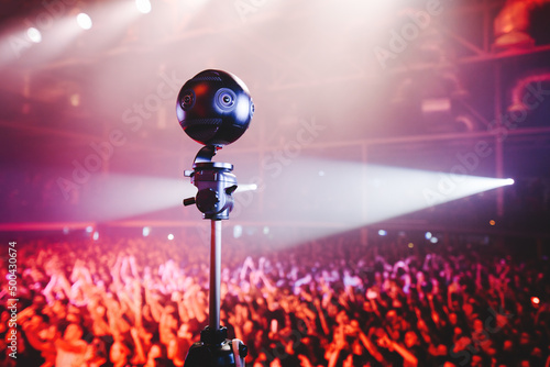Professional 360 camera at music concert on a tripod recording performance on video. silhouettes of crowd in front of bright stage lights.