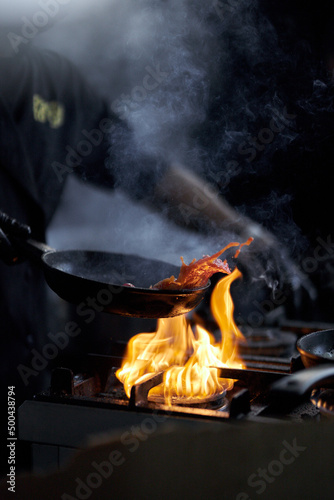 The cook prepares the sauce in a frying pan with fire. chef cooking, food processing with flame in frying pan on dark background, restaurant service concept, impressive cooking. Cooking with fire.