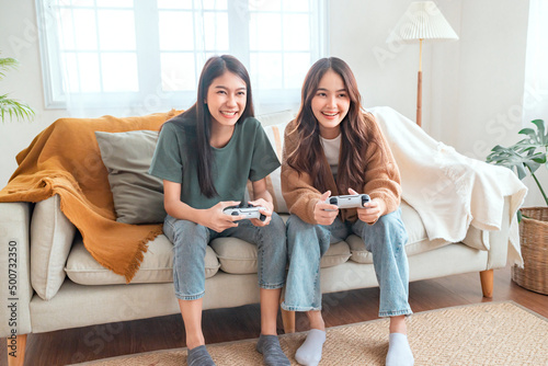 Two happy asian roommates sitting on couch in living room at home enjoy and excited holding console playing game together.