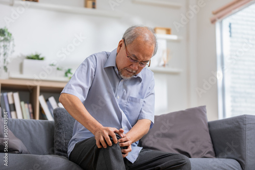 Asian old man with his knee joint pain in sofa, pain in the elderly, health care, elderly care. Elderly man having a knee pain and sitting down. Grandfather with knee pain.