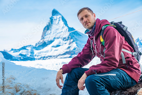Portrait of smiling tourist sitting on rock. Famous snowcapped matterhorn mountain against sky. Young male is relaxing in alpine region.