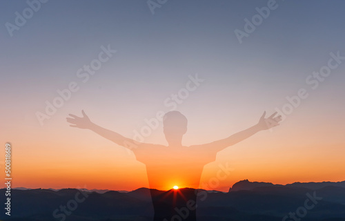 Religious man with arms raised thanking and praying to God, with the sunset in the mountains in the background.