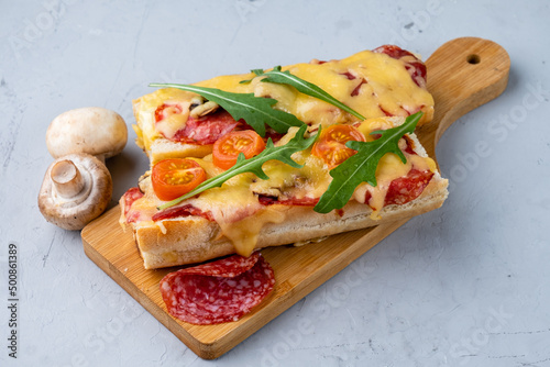 Homemade pizza sandwich, pizza cooked on a baguette with cheese, pepperoni sausages and mushrooms, served on a wooden board gray background.