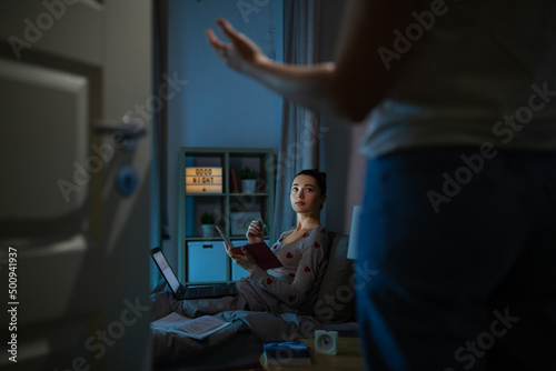 education, technology and people concept - teenage student girl with notebook and laptop computer learning in bed at home at night and mother entering room