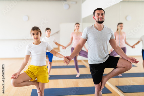 Young adult man practicing yoga in pair with teen daughter, performing double tree pose during family workout at gym
