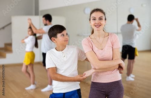Smiling preteen boy learning dynamic boogie-woogie in pair with his young mother in dance studio during family class..