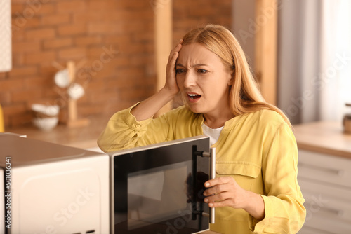 Worried mature woman near damaged microwave oven in kitchen