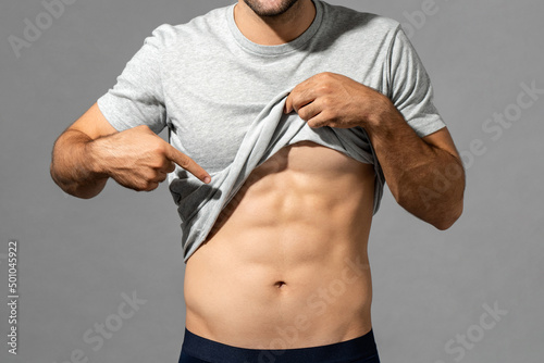 Muscular male model lifting up t-shirt to show abs while standing on gray isolated background in studio