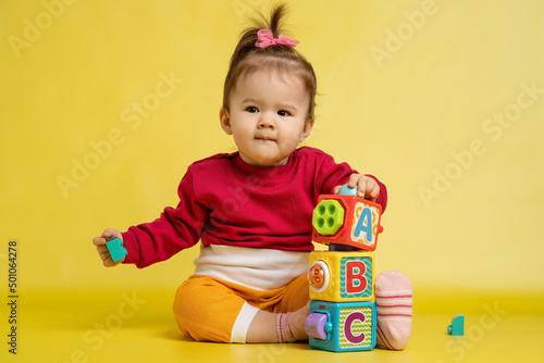 Girl 1 year old, playing with educational cubes with the English alphabet. The girl is sitting on the floor on a yellow background.