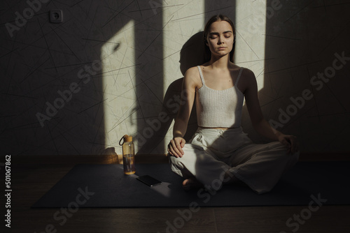Quiet woman with closed eyes sitting in Lotus pose on mat and doing yoga while meditating and practicing mindfulness in room with sunlight 