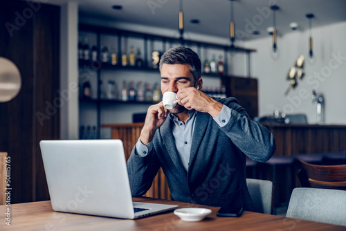 A businessman is sipping his coffee while looking at the laptop. 