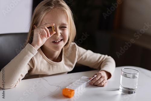 Little Caucasian Girl in pajamas taking natural vitamin omega-3 from plastic box on kitchen at home early morning. Adorable child Takes supplements.
