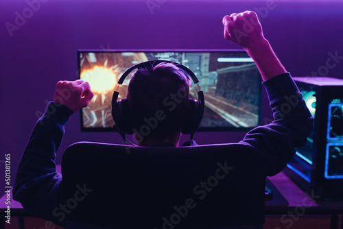Back view of pro gamer wearing headset celebrating victory in online internet esport tournament, makes yes hands gesture, looking at monitor. Neon coloured dark room. Cyber sport and e-sport concept