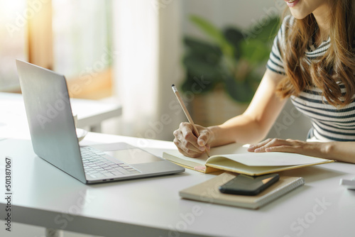 Close up image of a woman writing and taking note on notebook with laptop in office.