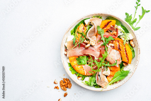 Summer salad bowl with sweet grilled peach, jamon, soft cheese, walnuts and fresh arugula on white kitchen table background, top view, negative space