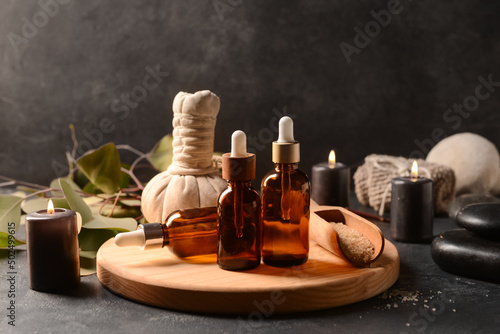 Wooden board with bottles of essential oil, sea salt, massage bag and burning candles on dark background