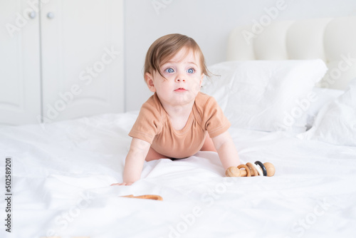baby girl with blue eyes in brown bodysuit playing with wooden toys on white bedding on bed