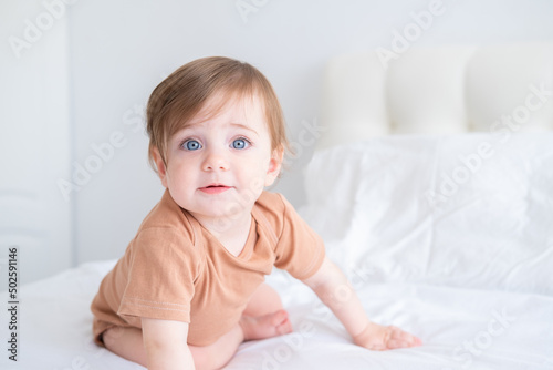 portrait of cute baby girl 10 months with blue eyes in brown bodysuit on white bedding on bed