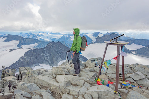 young woman alpinist standing on the top of the Galdhopiggen mountain summit in Norway. White and grey snow and ice, blue sky with clouds, rocks and dark mountains around