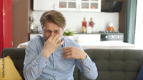 Unhappy, sullen man sitting in his armchair at home. Man bothered by bad smell and covering his nose.The young man feels an unpleasant odor.