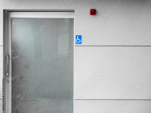Small blue disable handicap toilet sign on the wall at outdoor restroom door with fire alarm. public disabled toilet with frosted glass door..